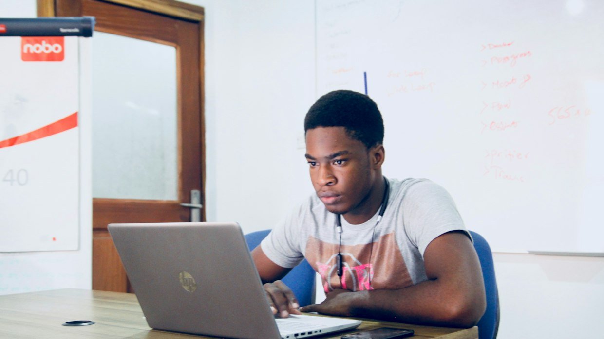 A student writing notes while a lecture plays on a laptop screen.